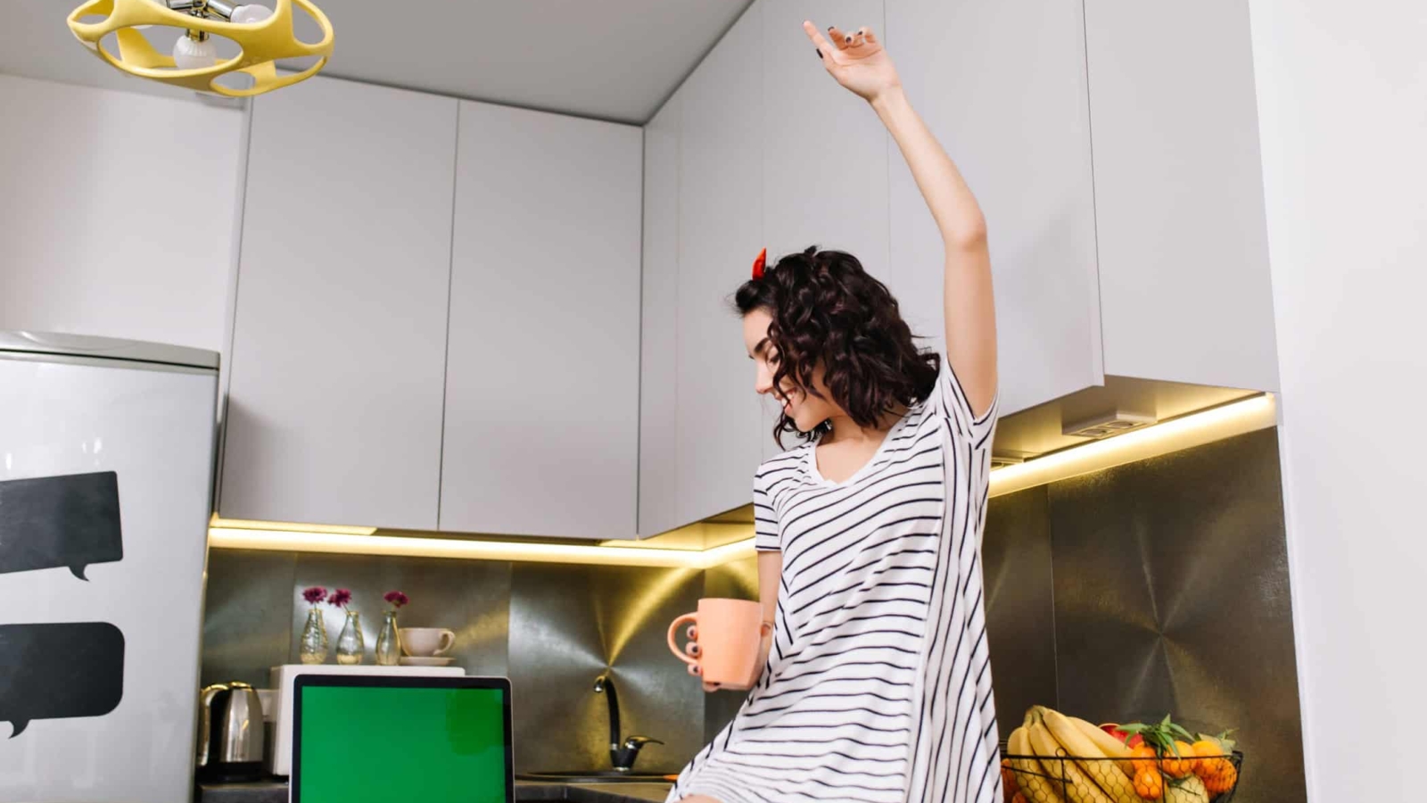 Happy morning in kitchen in modern apartment of joyful young woman in dress having fun on table. Laptop with green screen, tea, brightful emotions, smiling, true emotions