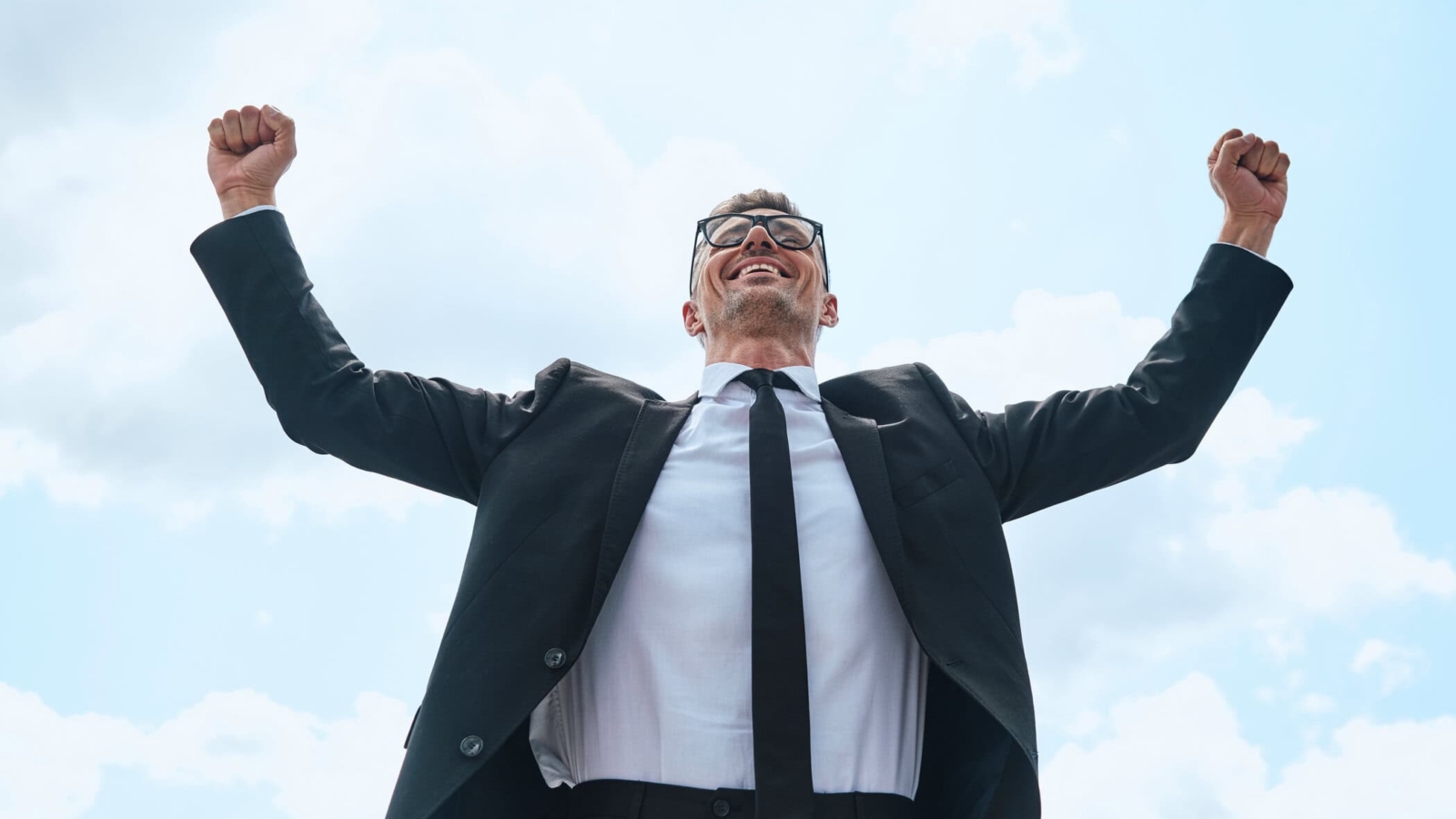 Happy mature man in full suit keeping arms raised while standing in front of blue sky outdoors