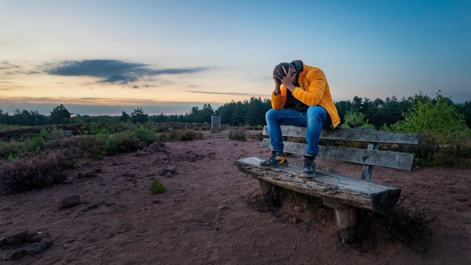 A sad older male clutching his head in his hands on an old wooden bench on a rural field